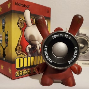 Könnte beinhalten: Rotes und schwarzes Dunny-Spielzeug mit einem Kameraobjektiv als Kopf. Das Spielzeug steht auf einer weißen Oberfläche. Die Schachtel für das Spielzeug befindet sich im Hintergrund und zeigt ein weißes Kaninchen mit einem Zylinder und den Worten "Kidrobot Dunny Side Show".