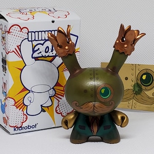 May include: A green and gold Dunny toy with a crown on each ear, a gold mustache, and a green suit. The toy is standing on a white surface. The toy is from the Kidrobot Dunny 2012 series.