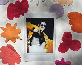 May include: A Polaroid photograph of a person wearing a yellow jacket, black shirt, and white trousers. The person is wearing sunglasses and has a belly button piercing. The photo is surrounded by colourful paper shapes.