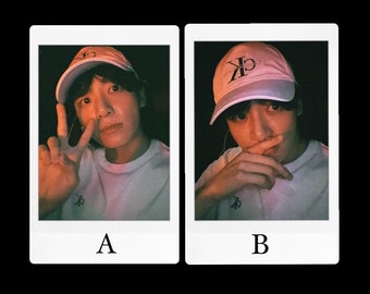 May include: Two Polaroid photos of a person wearing a white baseball cap with a black logo. The person is wearing a white t-shirt. In the first photo, the person is making a peace sign with their hand. In the second photo, the person is covering their mouth with their hand.