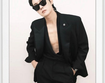 May include: A man wearing a black suit jacket with a deep V-neck, black trousers, and black sunglasses. He is standing in front of a white background.