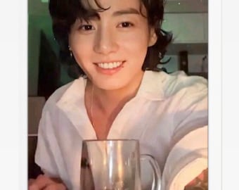 May include: A man with dark hair is smiling and holding a clear glass mug. The mug is filled with a light-coloured beverage. The background is blurry and features a green and white pattern.