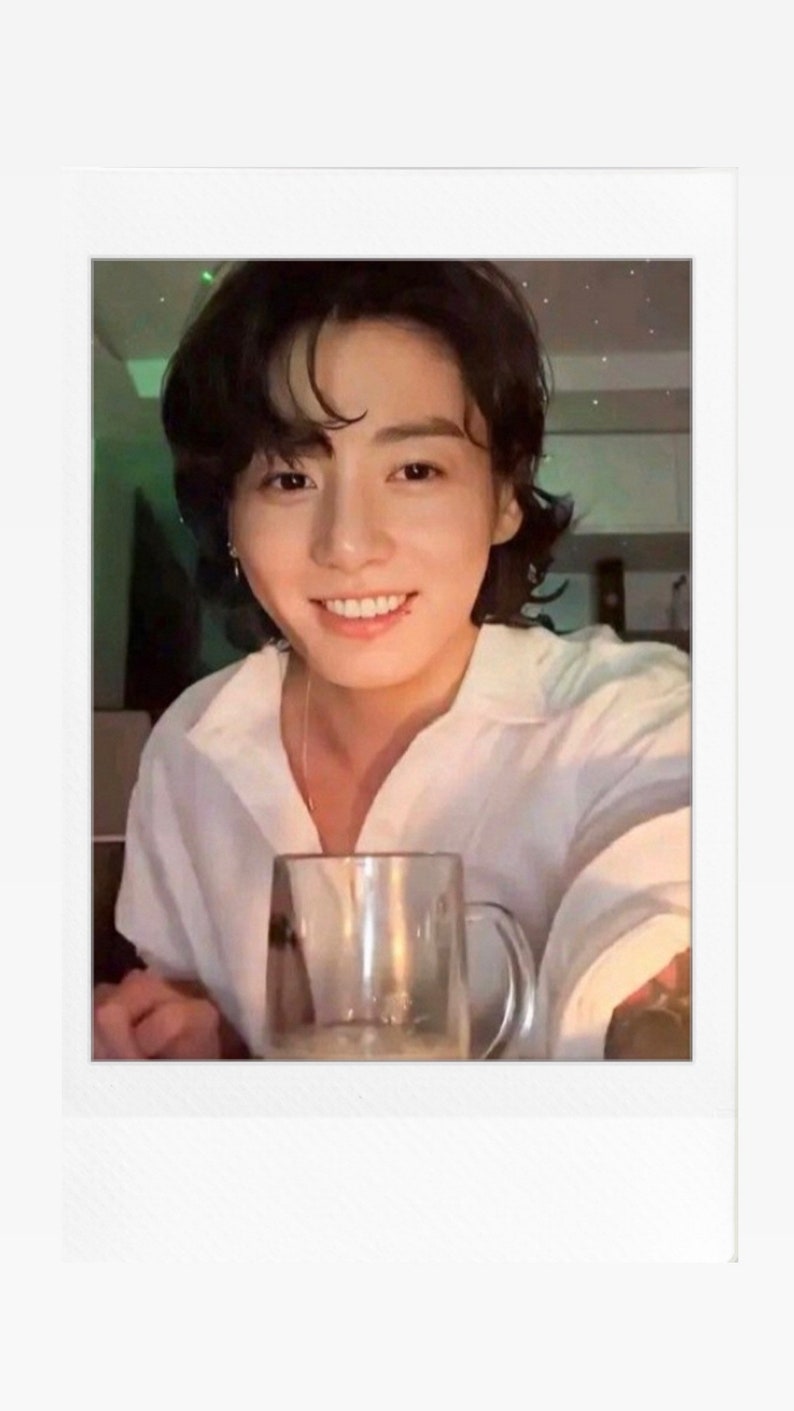 May include: A man with dark hair is smiling and holding a clear glass mug. The mug is filled with a light-coloured beverage. The background is blurry and features a green and white pattern.