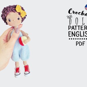 Puede incluir: Patrón de muñeca de crochet en inglés, formato PDF. La muñeca es de piel morena con el pelo rizado marrón, lleva un mono azul, zapatos rojos y sostiene una manzana roja y amarilla.