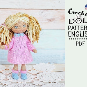 May include: Crochet doll pattern in English, PDF format. The pattern features a doll with blonde hair, a pink dress with white dots, and blue shoes. The doll is standing on a white crocheted doily.