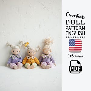 Op de afbeelding: Drie handgemaakte gehaakte poppen met garen haar en bloemenkragen. De poppen dragen truien in paars en geel, met bijpassende broeken. De afbeelding bevat de tekst "Crochet Doll Pattern English" en een pictogram van de Amerikaanse vlag.