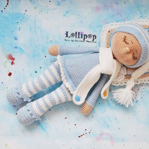May include: A crocheted doll wearing a blue and white striped outfit and a white hat, sleeping on a pillow with a crocheted border. The doll is lying on a blue background with white and coloured dots.