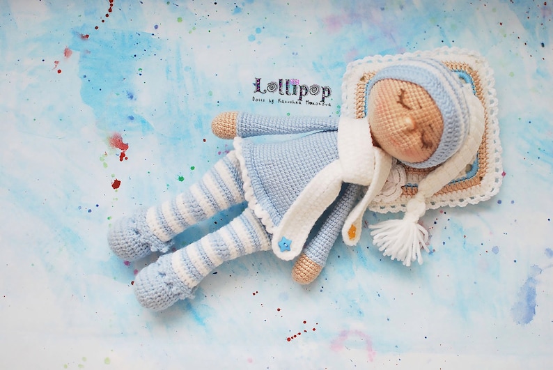 May include: A crocheted doll wearing a blue and white striped outfit and a white hat, sleeping on a pillow with a crocheted border. The doll is lying on a blue background with white and coloured dots.