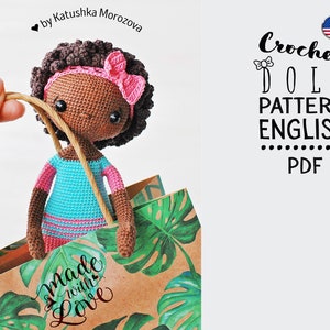 Puede incluir: Patrón de muñeca de crochet en inglés, formato PDF. El patrón presenta una muñeca de piel morena con cabello castaño, un lazo rosa y un vestido azul y rosa. La muñeca está en una bolsa de papel marrón con hojas verdes.