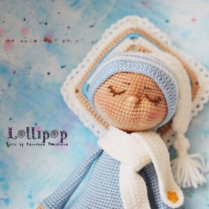 May include: A crocheted doll wearing a blue hat and scarf, lying on a crocheted blanket. The doll has closed eyes and a light brown complexion. The doll is labelled "Lollipop Dolls by Katusha Moiseeva".