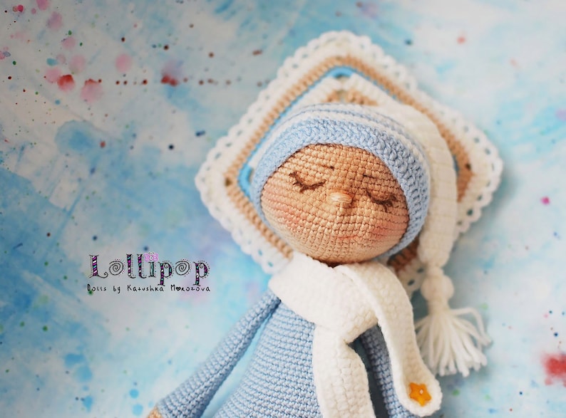 May include: A crocheted doll wearing a blue hat and scarf, lying on a crocheted blanket. The doll has closed eyes and a light brown complexion. The doll is labelled "Lollipop Dolls by Katusha Moiseeva".