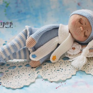 May include: A crocheted baby doll wearing a blue and white striped outfit, a white hat, and a white scarf. The doll is lying on a white crocheted blanket with a white tassel.