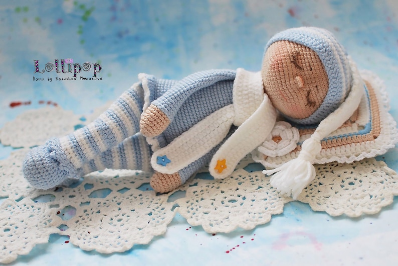 May include: A crocheted baby doll wearing a blue and white striped outfit, a white hat, and a white scarf. The doll is lying on a white crocheted blanket with a white tassel.