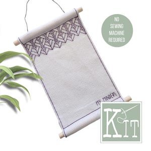 Pode incluir: Kit de ponto cruz branco com um design geométrico em fio roxo escuro. O kit é montado em cavilhas de madeira e inclui o texto "MY MINDERS" e "KIT". Um círculo verde diz "NO SEWING MACHINE REQUIRED".