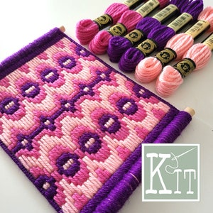 Pode incluir: Uma tapeçaria de ponto de agulha rosa e roxa com um padrão geométrico. A tapeçaria está em um rolo de madeira e vem com um kit de fio de bordado rosa e roxo.