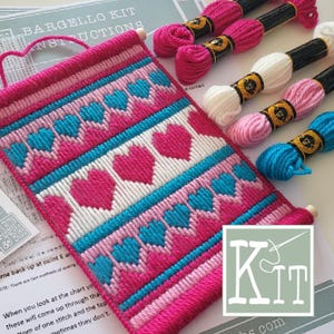 Pode incluir: Um projeto de bordado Bargello com um padrão de corações repetidos em tons de rosa choque, turquesa e branco. O kit inclui uma folha de instruções impressa e cinco meadas de linha de bordar em tons de rosa, branco e azul.