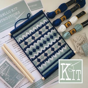 Pode incluir: Um kit de ponto de agulha com uma tapeçaria parcialmente bordada com um padrão geométrico em azul-marinho, azul-claro e branco. Inclui duas varetas de madeira, quatro novelos de fio em azul-marinho, branco e azul-claro e instruções.