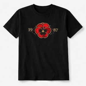 Op de afbeelding: Zwart T-shirt met een rood en goud bloemdesign en de gouden cijfers "1987". De bloem heeft een geborduurd uiterlijk. Het T-shirt is een klassieke ronde hals.
