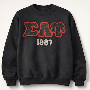 May include: Black crewneck sweatshirt with the letters "ΣΑΠ" in a red outline and the year "1987" in white. The sweatshirt is a solid black color.