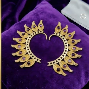 May include: A pair of gold-colored earrings with a leaf design and purple gemstone accents. The earrings are arranged on a purple velvet surface, showcasing their intricate details and elegant style.