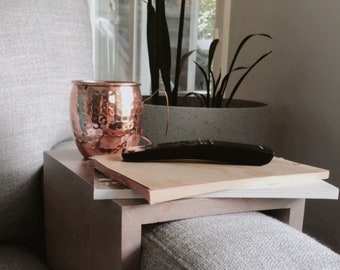 May include: A copper Moscow Mule mug and a black remote rest on a wooden side table attached to a gray upholstered sofa. A potted plant is visible in the background near a window.
