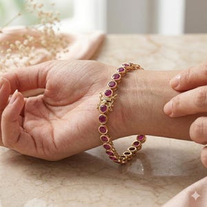 May include: A gold-toned bracelet featuring a series of round, bezel-set vibrant pinkish-red stones linked together. The piece includes a secure box clasp mechanism with a safety latch, creating a continuous circular design around the wrist.