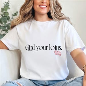 Puede incluir: Camiseta blanca con la frase "Gird your loins" en fuente negra. Debajo, en rojo, el texto "The Devil Wears Prada". Una camiseta casual y cómoda.