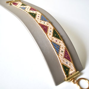 K&ouml;nnte beinhalten: Ein Armband aus Perlen mit einem geometrischen Muster in Rosa-, Blau-, Gr&uuml;n- und Goldt&ouml;nen. Das Armband ist aus kleinen Perlen gefertigt und hat einen goldenen Verschluss.