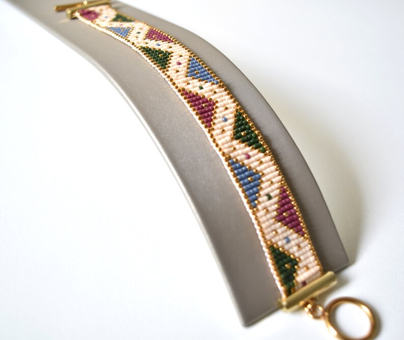 K&ouml;nnte beinhalten: Ein Armband aus Perlen mit einem geometrischen Muster in Rosa-, Blau-, Gr&uuml;n- und Goldt&ouml;nen. Das Armband ist aus kleinen Perlen gefertigt und hat einen goldenen Verschluss.