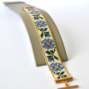 May include: A white beaded bracelet with a blue and gold floral pattern. The bracelet has a gold clasp.