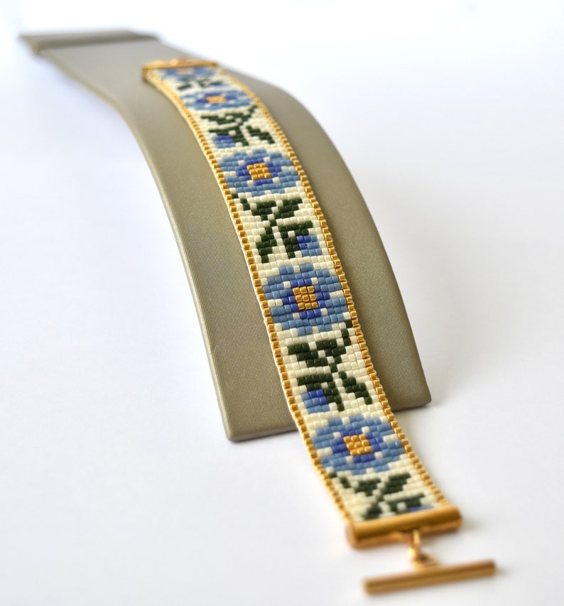 May include: A white beaded bracelet with a blue and gold floral pattern. The bracelet has a gold clasp.