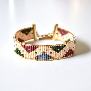 K&ouml;nnte beinhalten: Ein Armband aus Perlen mit einem geometrischen Muster in Gr&uuml;n-, Blau-, Rosa- und Wei&szlig;t&ouml;nen. Das Armband hat einen goldenen Verschluss.