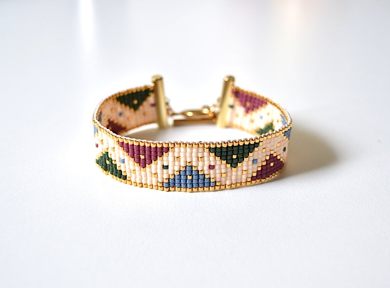 K&ouml;nnte beinhalten: Ein Armband aus Perlen mit einem geometrischen Muster in Gr&uuml;n-, Blau-, Rosa- und Wei&szlig;t&ouml;nen. Das Armband hat einen goldenen Verschluss.