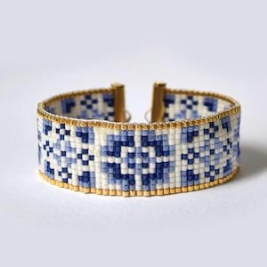 Puede incluir: Un brazalete de cuentas azul y blanco con un patrón geométrico. El brazalete está hecho con cuentas pequeñas y cuadradas y tiene un cierre dorado.