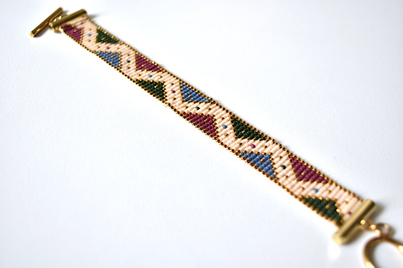 K&ouml;nnte beinhalten: Ein Armband aus Perlen mit einem geometrischen Muster in Rosa-, Blau-, Gr&uuml;n- und Goldt&ouml;nen. Das Armband hat einen goldenen Verschluss.