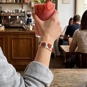 Peut inclure: Une main tenant un verre de smoothie rose garni de fraises et de menthe. Un bracelet de perles avec un motif de fraises est porté au poignet. En arrière-plan, un café avec des étagères et des tables en bois.