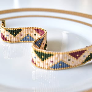 K&ouml;nnte beinhalten: Ein Armband aus Perlen mit einem geometrischen Muster in Beige-, Gr&uuml;n-, Blau- und Burgunder-T&ouml;nen. Das Armband hat einen goldenen Verschluss.