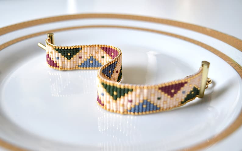 K&ouml;nnte beinhalten: Ein Armband aus Perlen mit einem geometrischen Muster in Beige-, Gr&uuml;n-, Blau- und Burgunder-T&ouml;nen. Das Armband hat einen goldenen Verschluss.