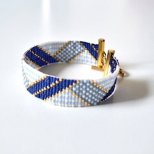 Könnte beinhalten: Ein weißes, blaues und goldenes Armband mit geometrischem Muster. Das Armband hat einen goldenen Verschluss.