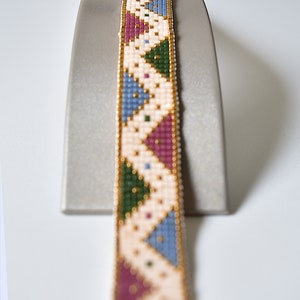 K&ouml;nnte beinhalten: Ein Armband aus Perlen mit einem geometrischen Muster in Rosa-, Blau-, Gr&uuml;n- und Wei&szlig;t&ouml;nen. Das Armband befindet sich auf einem Pr&auml;sentationsst&auml;nder.