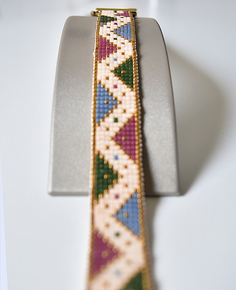 K&ouml;nnte beinhalten: Ein Armband aus Perlen mit einem geometrischen Muster in Rosa-, Blau-, Gr&uuml;n- und Wei&szlig;t&ouml;nen. Das Armband befindet sich auf einem Pr&auml;sentationsst&auml;nder.
