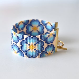 Op de afbeelding: Een kralen armband met een bloemenpatroon in tinten blauw, wit en geel. De armband heeft een gouden sluiting.