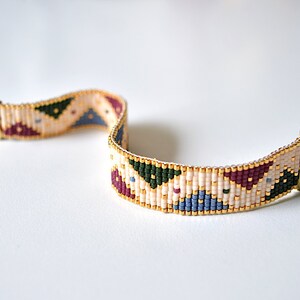 K&ouml;nnte beinhalten: Ein Armband aus Perlen mit einem geometrischen Muster in Beige-, Burgunder-, Gr&uuml;n- und Blaut&ouml;nen. Das Armband hat goldfarbene Verschl&uuml;sse.