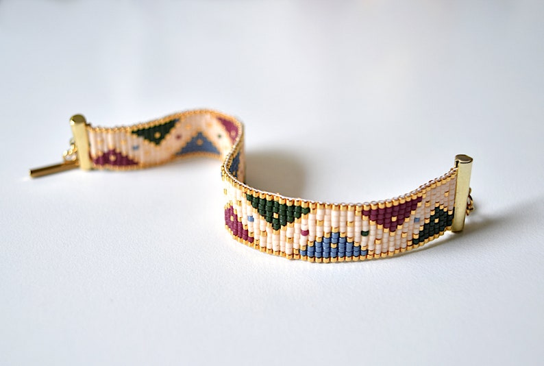 K&ouml;nnte beinhalten: Ein Armband aus Perlen mit einem geometrischen Muster in Beige-, Burgunder-, Gr&uuml;n- und Blaut&ouml;nen. Das Armband hat goldfarbene Verschl&uuml;sse.