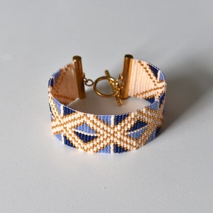 May include: A beaded bracelet with a geometric pattern in shades of blue, white, and gold. The bracelet has a gold clasp.