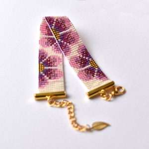 Może przedstawiać: Bransoletka z koralików w kolorze różowym i białym z kwiatowym wzorem i złotymi akcentami. Bransoletka ma złoty łańcuszek i zapięcie.