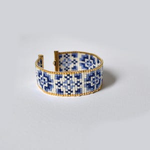 Puede incluir: Un brazalete de cuentas blancas y azules con un patrón geométrico. El brazalete tiene un cierre dorado.