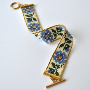 May include: A white beaded bracelet with a blue floral pattern and gold accents. The bracelet has a toggle clasp.