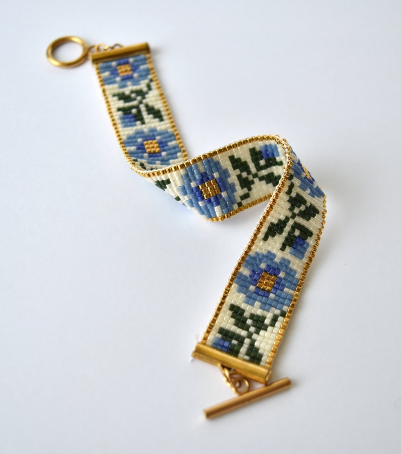 May include: A white beaded bracelet with a blue floral pattern and gold accents. The bracelet has a toggle clasp.