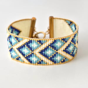 May include: A white beaded bracelet with a geometric pattern in shades of blue and gold. The bracelet has a gold clasp.